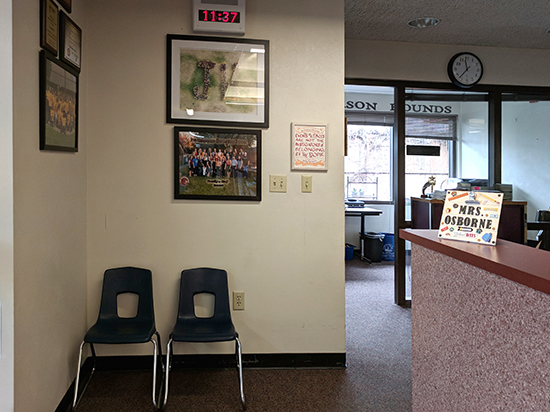 The certificate installed on a wall with a group photos. There are two child-size chairs in front of the wall. To the right is a counter where a handmade sign reads Mrs. Osborne. In the background is the principal's office.