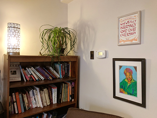 The certificate installed above a drawing of Maya Angelou, which is above a comfy chair. It's to the right of a softy-lit lamp  and a houseplant on top of a bookshelf.