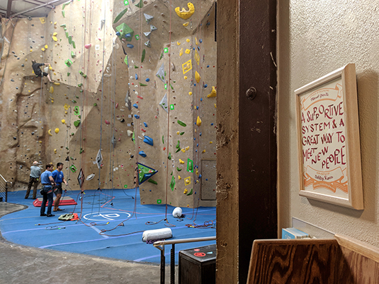 Certificate hanging on a wall on the right of the image. In the distance is an indoor rock climbing gym. A few climbers are on the ground, while one climber is on the wall.