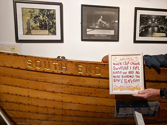 Hand holding a certificate in front of a boat with the words South End. Behind the boat are black and white photos of rowers on the wall.