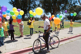 A dark-skinned man on a bicycle stopped to recieve a ballon from a row of kids from a Chinatown youth program.
