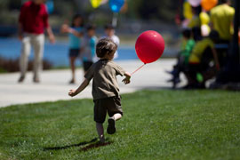 a young toddler runs on an expanse of lawn. His face is turned to the red balloon tied to his wrist. In the backgound are project team members wearing yellow t-shirts.