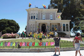 A large group of people are setting up on the front lawn of the Camron-Stanford House, a Victorian house on Lake Merritt, Oakland, CA. The team members are easily identified by their yellow t-shirts. There is also signage of the title, The Great Ballon Giveaway. Each letter was cut from poster board and affixed to the iron fence surrounding the lawn, facing a busy intersection.