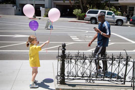 A primary-school aged girl extends a balloon to a stranger who is approaching on the sidewalk. 