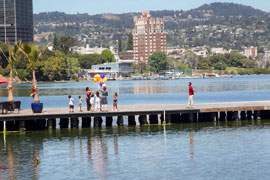 A photo of the lake. In the middle distance, on a wooden dock jutting into the lake, are a group of team-members sorting out a bunch of orange and purple balloons while strangers look on. In the background are treess in the park and buildings in neighborhing Adams point.