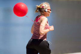 A woman jogger runs. She is wearing sunglasses and headphones. A red balloon trails behind her. In the background is Lake Merritt.