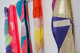 close up of the sashes hanging on a dowel in the gallery. each sash has a embroidered label that reads 'Irrational Exuberance (Asst Colors) Christine Wong Yap' in gold text on white background.