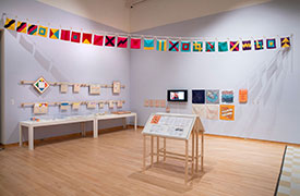 installation view of an art exhibit with semaphore flags above, cases with ephmera displays, and drawings and prints on the walls.