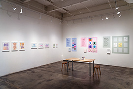 gallery installation with prints on the left wall, and different colored activity areas on the back wall. 