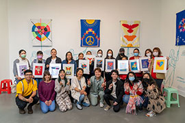 A group photo of many Chinese and Latinx women holding miniature framed versions of their banners, along with project team members, in the gallery in front of large banners.