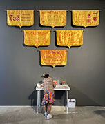 a young girl writes at a desk beneath an installation of six yellow banners