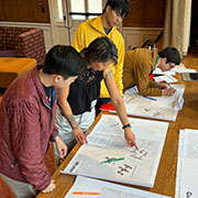 An Asian American woman points at a large printout of a map of the Wisteria neighborhood while two students  look on. A third student writes on another map in the background.