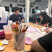 Students paint at a table, with a cup of brushes in the foreground.