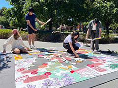 Students confer with each other while holding puzzle pieces in their hands while other puzzle pieces are assembled on a grey mat on the ground. Trees are in the background.