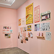 A pink gallery wall features many items, including a large banner for the Persian Poetry Forum of Los Angeles, vinyl map of places of belonging in the San Francisco Bay Area, and framed works. Books and zines sit on shelves for perusal.