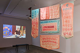 Banners using pink and blue colors feature hand-lettered calligraphy with quotes in different languages. The banners hang in the middle of the space. with a video projection on the back wall of a woman in front of another banner.