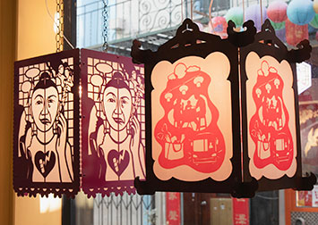 two lanterns hanging in a window. One is purple with a woman's sad/happy face. One is wood with a red papercut of scenes in Chinatown